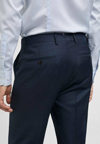 Pantalon militaire ajusté en tissu texturé, avec deux poches arrière à fermeture boutonnée. Une chemise habillée bleu clair est partiellement visible.