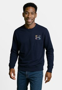 Marineblauwe sweatshirt van een zachte stof met een borstprint met veelkleurige accenten en geribbelde manchetten en zoom. Draagt een blauwe jeans.