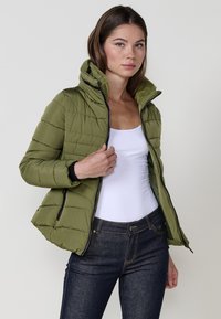 Chaqueta acolchada de color verde oliva con un alto cuello, cierre frontal con cremallera y textura acolchada. Presenta detalles en negro y se combina con jeans oscuros ajustados.