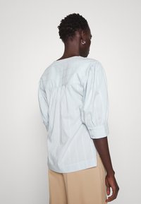 Lyseblå stripete bluse med en avslappet passform, puffede ermer og knappede mansjetter. Har en buet kant og detaljering på bakstykket.