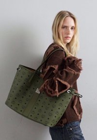 Blonde woman wearing a brown distressed sweater and jeans, carrying a large green patterned tote bag over her shoulder against a plain wall.