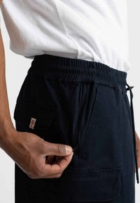 Navy blauwe elastische broek met zijzakken, vervaardigd uit zachte stof. Voorzien van een trekkoord en een label met de tekst "REVOL." De textuur oogt glad.