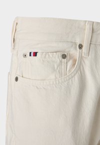 Pantalones de mezclilla beige con bolsillos delanteros y traseros. Las características notables incluyen herrajes de metal y un pequeño emblema tricolor en el bolsillo trasero.