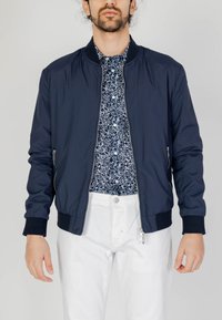 Giacca bomber navy con tasche con zip, polsini e colletto a costine. Indossata sopra una camicia bianca e navy a fantasia, abbinata a pantaloni bianchi.