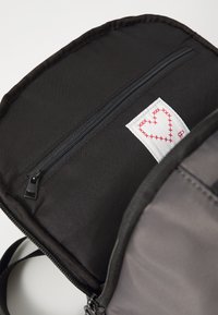 Černý látkový batoh s kapsou na zip na přední straně. Obsahuje bílou nálepku s vyšitým červeným srdíčkem. Hladký povrch, měkké okraje.