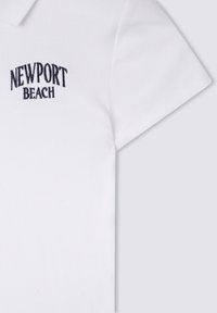 Polo bianco realizzato in tessuto a coste. Presenta "NEWPORT BEACH" ricamato in blu navy sul petto. Maniche corte e design del colletto classico.