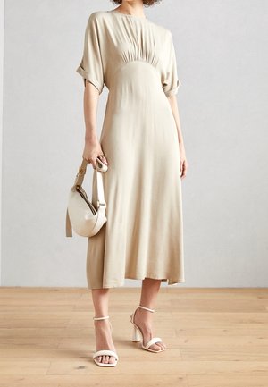 Femme portant une robe midi beige avec un détail froncé, tenant un sac à main beige assorti et portant des sandales blanches à talons sur un sol en bois.