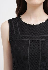Ärmlös svart klänning i texturerad vadderad tyg, med spetspaneler och en rund halsringning dekorerad med små metallnitar.