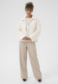 Woman with curly hair wearing hoop earrings, a white fuzzy jacket over a lace top, beige wide-leg pants, and burgundy pointed shoes.