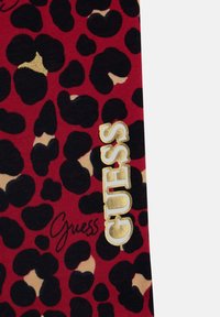Red fabric featuring a leopard print pattern in black and beige shades, with "GUESS" embroidered in gold and a signature in black.
