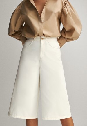 Blouse beige à manches bouffantes rentrée dans un short blanc taille haute, jambes larges et longueur au genou, avec poches avant et fermeture par bouton.
