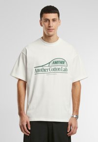 Tricou din bumbac alb, supradimensionat, cu text grafic verde, având inscripția "Another Cotton Lab" și un design cu o rachetă de tenis. Maneci scurte, gât rotund.