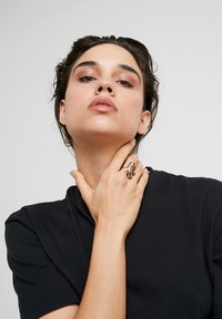 Gouden ring met ingewikkelde details, gedragen aan de rechterhand. Model draagt een zwart T-shirt en poseert met de hand bij de hals.