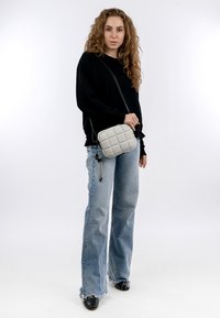 Borsa a tracolla grigia trapuntata con una tracolla nera, indossata sopra un maglione nero e jeans chiari strappati, su uno sfondo bianco.