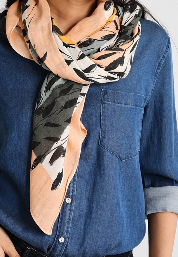 Femme portant une écharpe à motifs couleur pêche clair et noir enroulée autour du cou, sur une chemise en denim bleu à boutons avec les manches retroussées.