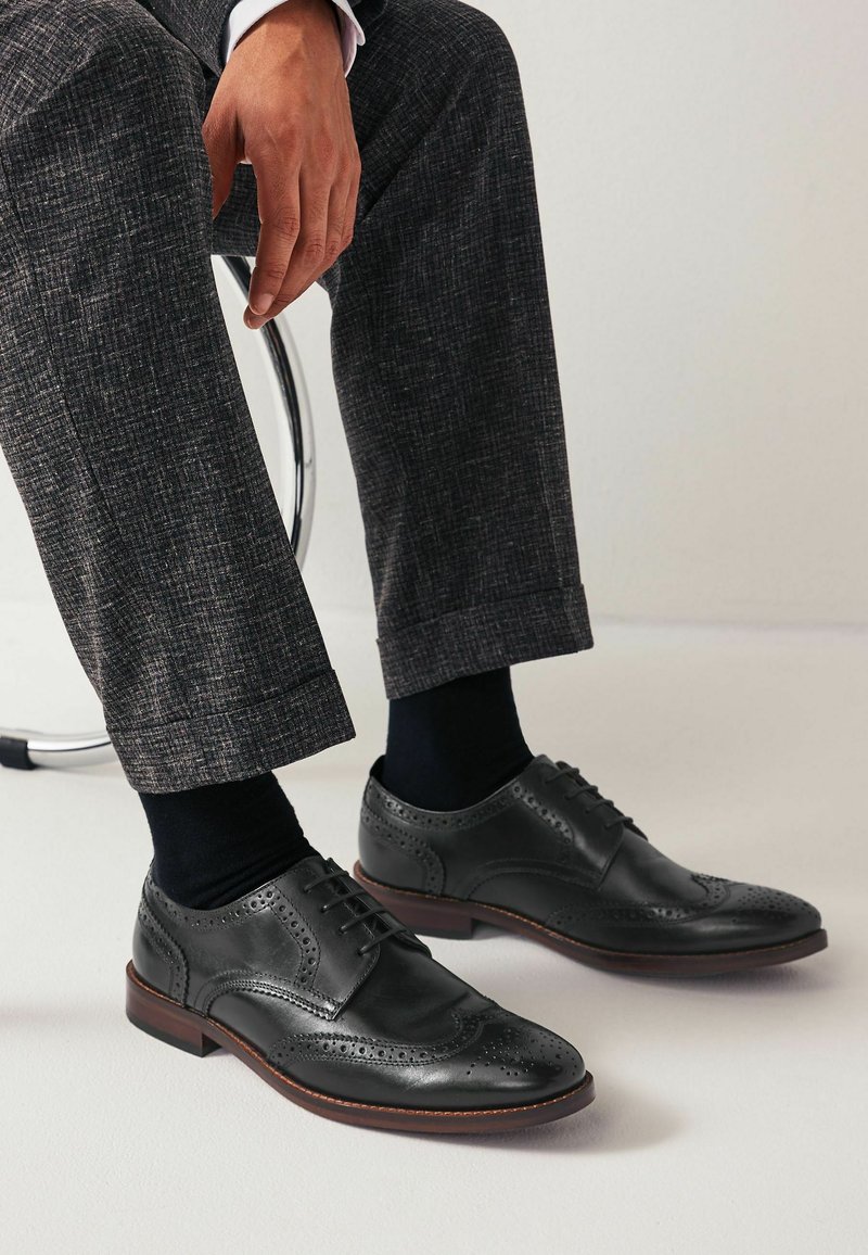 Zapatos de vestir de cuero negro con diseño de puntera alada, detalles de brogue y puntera redonda. Acompañados de calcetines de vestir oscuros y pantalones estampados.