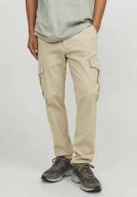 Khaki Cargo-Hosen mit mehreren Seitentaschen, aus Baumwolle, entspannte Passform und kombiniert mit grauen Sneakers. Das Shirt ist hellgrün und im Crew-Neck-Stil.