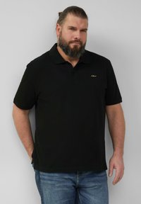 Zwarte polo met kraag, korte mouwen en een subtiel gouden logo op de linkerborst. Gekleed met een blauwe spijkerbroek. Glad stoftextuur.