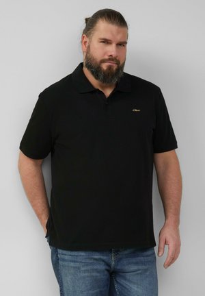 Zwarte polo met kraag, korte mouwen en een subtiel gouden logo op de linkerborst. Gekleed met een blauwe spijkerbroek. Glad stoftextuur.