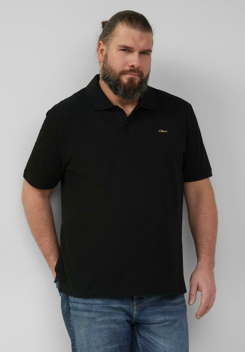 Zwarte polo met kraag, korte mouwen en een subtiel gouden logo op de linkerborst. Gekleed met een blauwe spijkerbroek. Glad stoftextuur.