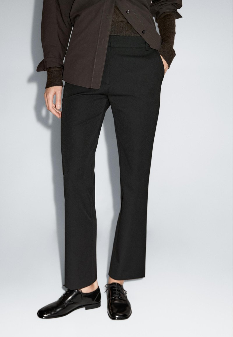 Zwarte tapered broek gemaakt van glanzende stof, met een middelhoge tailleband en zijzakken, gecombineerd met glanzende zwarte leren schoenen.