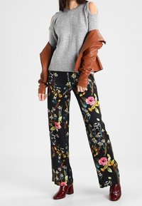 Grauer Cold-Shoulder-Pullover kombiniert mit schwarzen, blumigen, weiten Hosen, die lebendige Farben und eine glänzende Textur aufweisen, ergänzt durch burgunderfarbene Ankle Boots.
