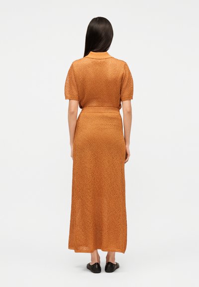 Hosszú, sötét hajú nő háttal áll, narancssárga, texturált rövid ujjú felsőt és hozzáillő, hosszú szoknyát visel fekete papuccsal.