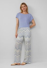 Lichtpaarse top met korte mouwen gecombineerd met wijd uitlopende broek met een zigzagpatroon in blauw, geel en wit. Het model draagt platte, geweven sandalen.