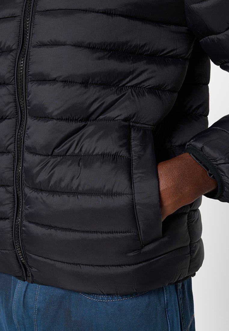 Gros plan sur une personne portant une veste matelassée noire avec une main dans la poche latérale et un pantalon en denim bleu.