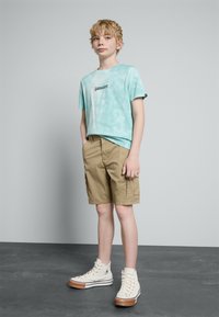 Φωτεινό μπλε tie-dye μπλουζάκι με μαύρο λογότυπο, χακί cargo σορτς και λευκά sneakers ψηλού σχεδίου με καφέ λεπτομέρειες, σε φόντο γκρίζου χρώματος.