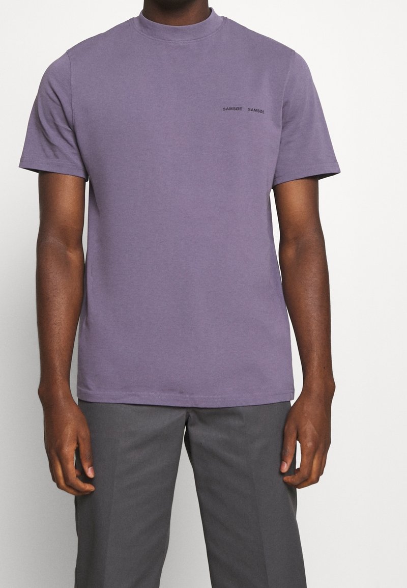 Homme portant un t-shirt simple violet à manches courtes avec un petit texte noir "SAMSOE SAMSOE" sur la poitrine gauche et un pantalon gris foncé.