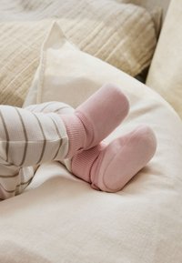 Chaussons pour bébé roses en tissu doux, avec des orteils arrondis et des bords côtelés, assortis à un pantalon rayé blanc et beige, reposant sur une surface à texture claire.