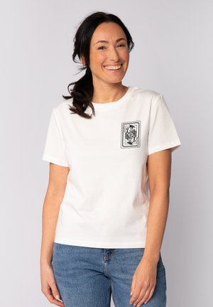 Lächelnde Frau mit dunklen Haaren im Pferdeschwanz trägt ein weißes T-Shirt mit einem schwarzen Spielkartendesign auf der Brust und blaue Jeans.