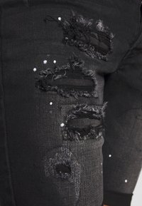 Jean en denim noir avec des patches usés aux bords effilochés et des éclaboussures blanches dispersées. Le matériau a une texture douce.
