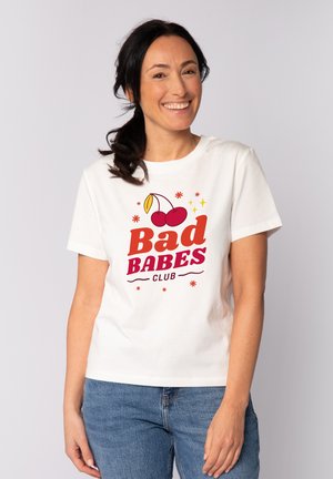 Vrouw met donker haar in een paardenstaart lacht en draagt een wit T-shirt met rode tekst "Bad Babes Club" en een kersenafbeelding, gecombineerd met een blauwe spijkerbroek.