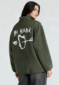 Veste en polaire oversize vert olive avec une texture douce. Présente un texte graphique blanc "Mi Amor" et un motif flèche au dos.