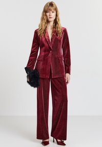 Completo in velluto rosso con blazer aderente e pantaloni a gamba larga, caratterizzato da un motivo sottile. Completo con clutch in pelliccia nera e tacchi.