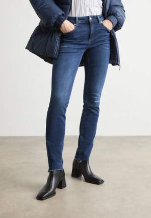 Personne portant un jean skinny bleu foncé légèrement usé, des bottines noires à talons, une chemise rayée et une doudoune bleu foncé les mains dans les poches.
