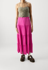 Top tipo halter en verde olivo con busto estructurado, combinado con una falda maxi rosa de múltiples capas. Unas sandalias de plataforma marrones completan el conjunto.