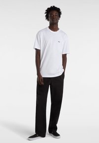 White cotton t-shirt with short sleeves, featuring a small logo on the chest. Paired with black, loose-fit trousers and black sneakers.