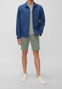 Un homme portant une veste bleue à boutons sur une chemise rayée, un short vert et des baskets blanches, se tenant contre un fond gris uni.