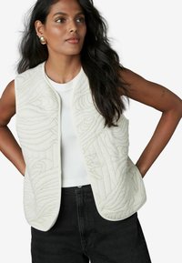 Gilet blanc matelassé avec des motifs ondulés élégants, devant ouvert, et une texture lisse, associé à un haut blanc ajusté et un jean noir.