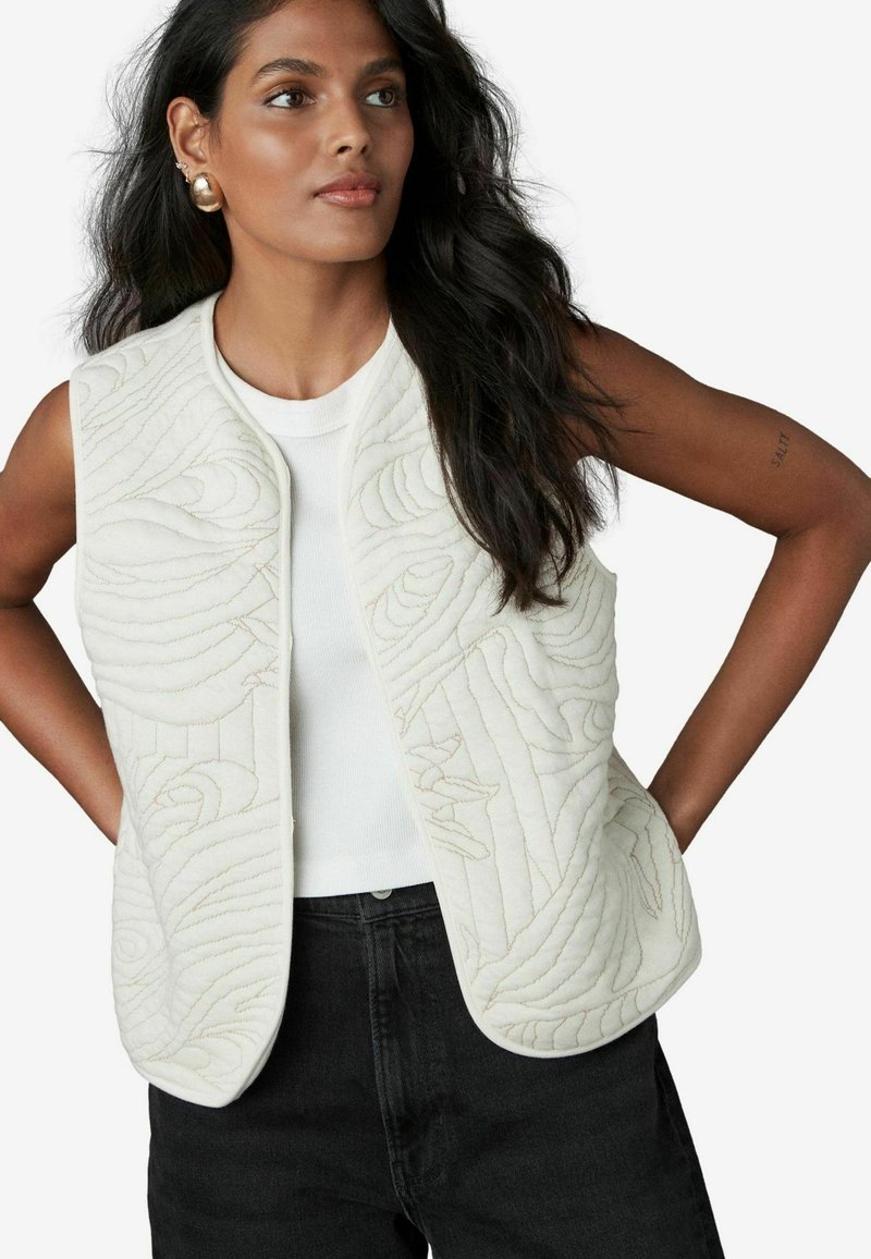 Gilet blanc matelassé avec des motifs ondulés élégants, devant ouvert, et une texture lisse, associé à un haut blanc ajusté et un jean noir.