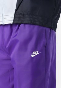 Prizor izbliza osobe koja nosi crnu i svijetloplavu jaknu preko ljubičastih sportskih hlača s bijelim Nike logom.