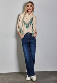 Maglietta a maniche lunghe beige chiaro, sciarpa verde e nera a fantasia, jeans a gamba larga blu scuro, sneaker bianche. Outfit semplice e casual.