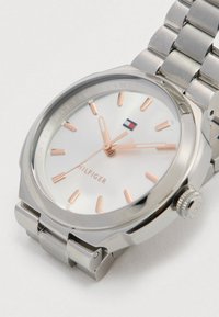 Ασημένιο ρολόι χειρός με ροζ χρυσούς δείκτες και δείκτες ωρών, λευκό καντράν, μεταλλικό λουρί και λογότυπο κοντά στην θέση 12.