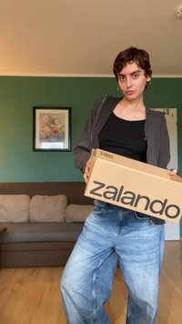 Kartónová krabice s nápisem "zalando" vytištěným černě, drží ji osoba v černém tílku a šedé zipové bundě, s modrými džíny.
