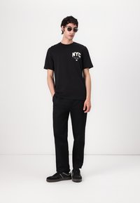 T-shirt noire en coton avec impression "NYC", associée à un pantalon noir et des baskets noires. Modèle portant des lunettes de soleil noires, se tenant devant un fond blanc.