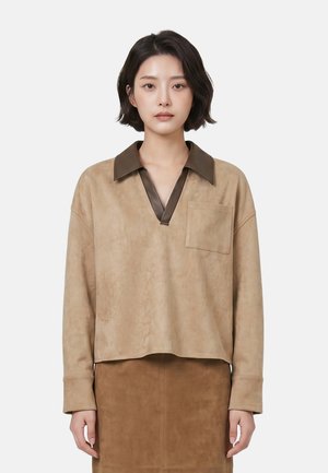 Jonge vrouw met kort donker haar draagt een beige top met lange mouwen en een donkerbruine kraag en een bijpassende bruine rok, staand tegen een witte achtergrond.