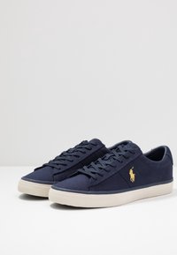 Granatowe płócienne trampki o teksturowanym wykończeniu, z beżową gumową podeszwą i złotymi akcentami logo po boku. Klasyczny design na sznurówki.